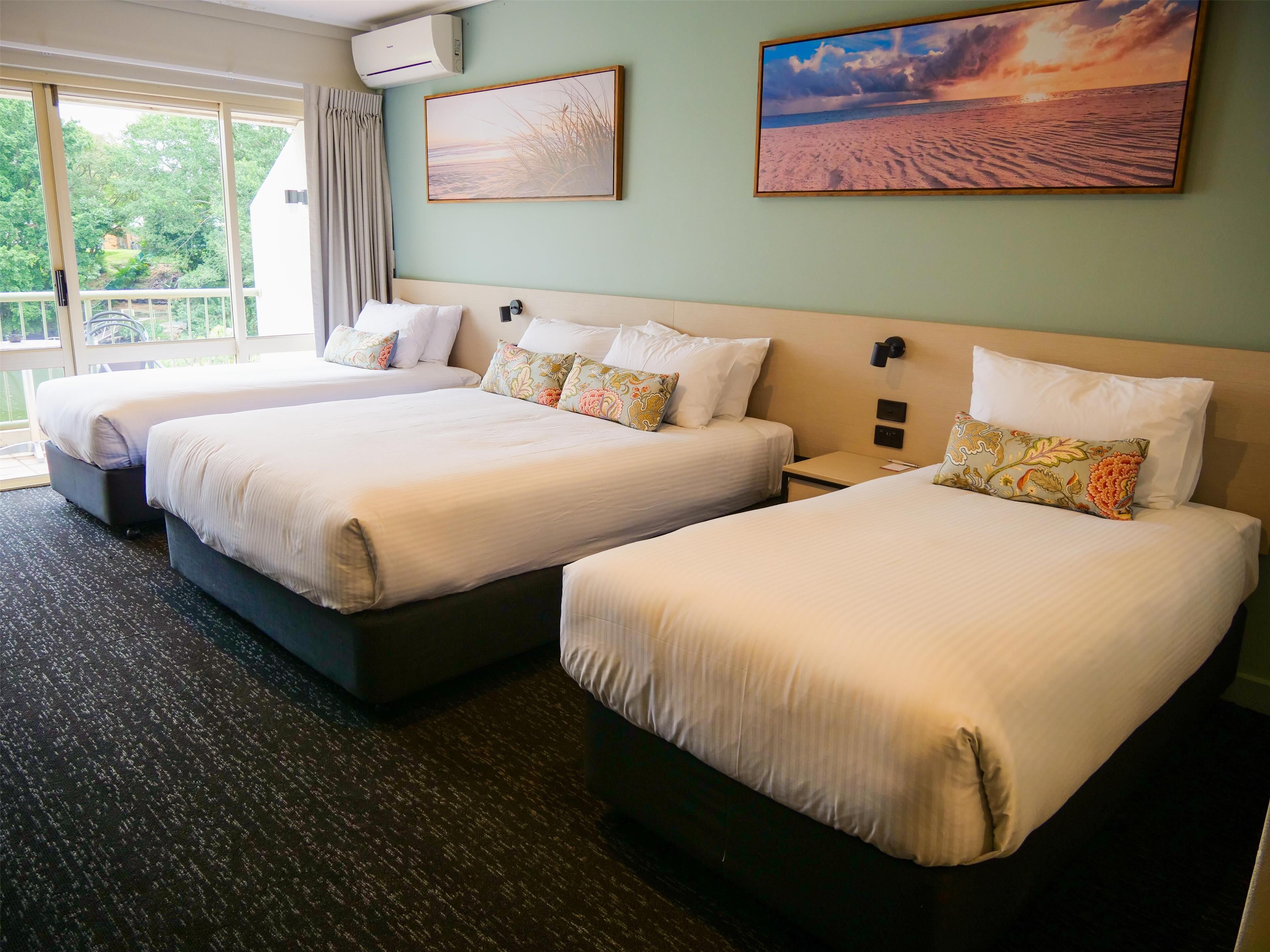 A hotel room with three beds, a large window with curtains, and two wall-mounted air conditioning units.