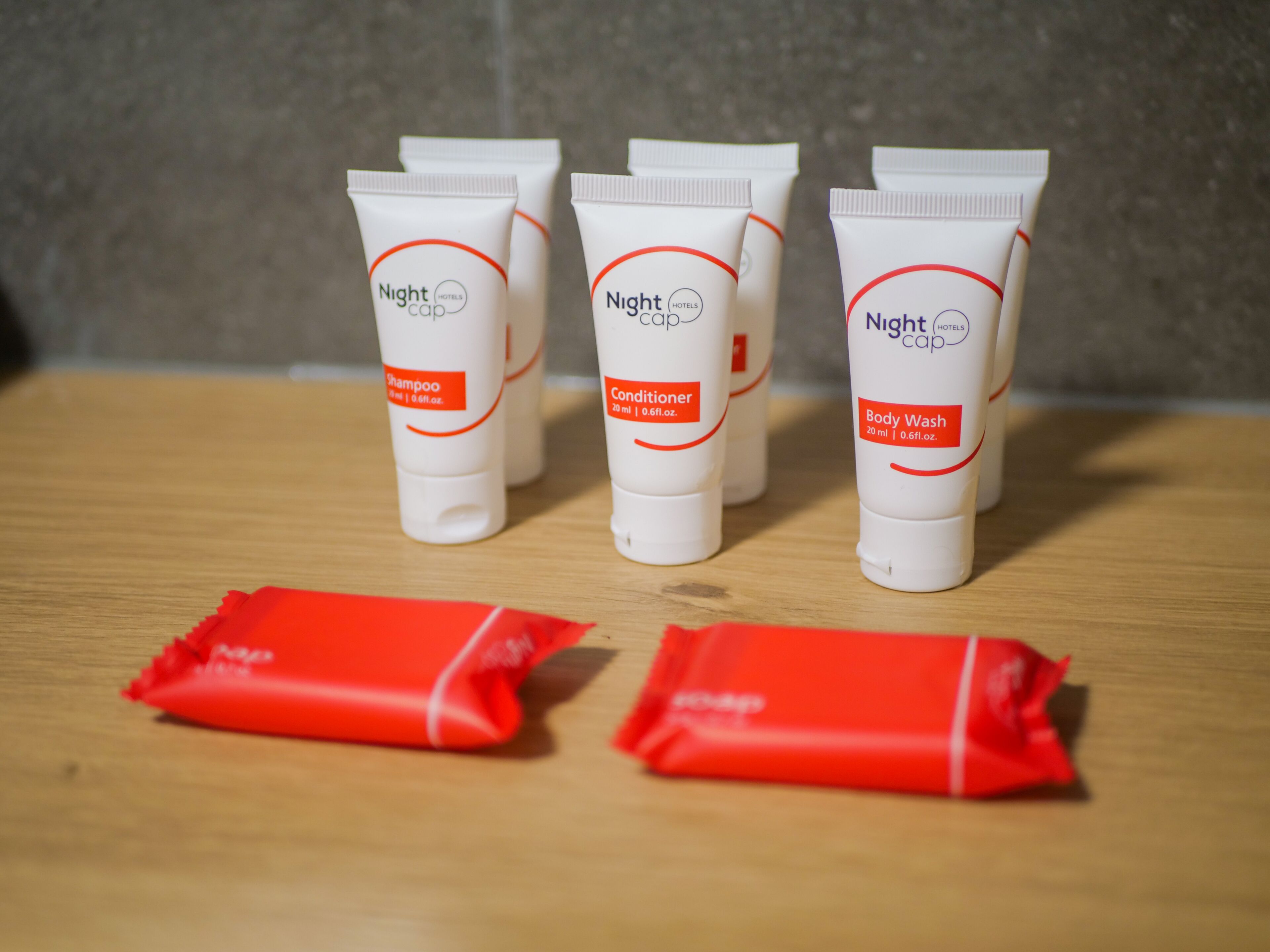 Four bottles of Night cap hotel toiletries and three red sachets on a wooden surface.