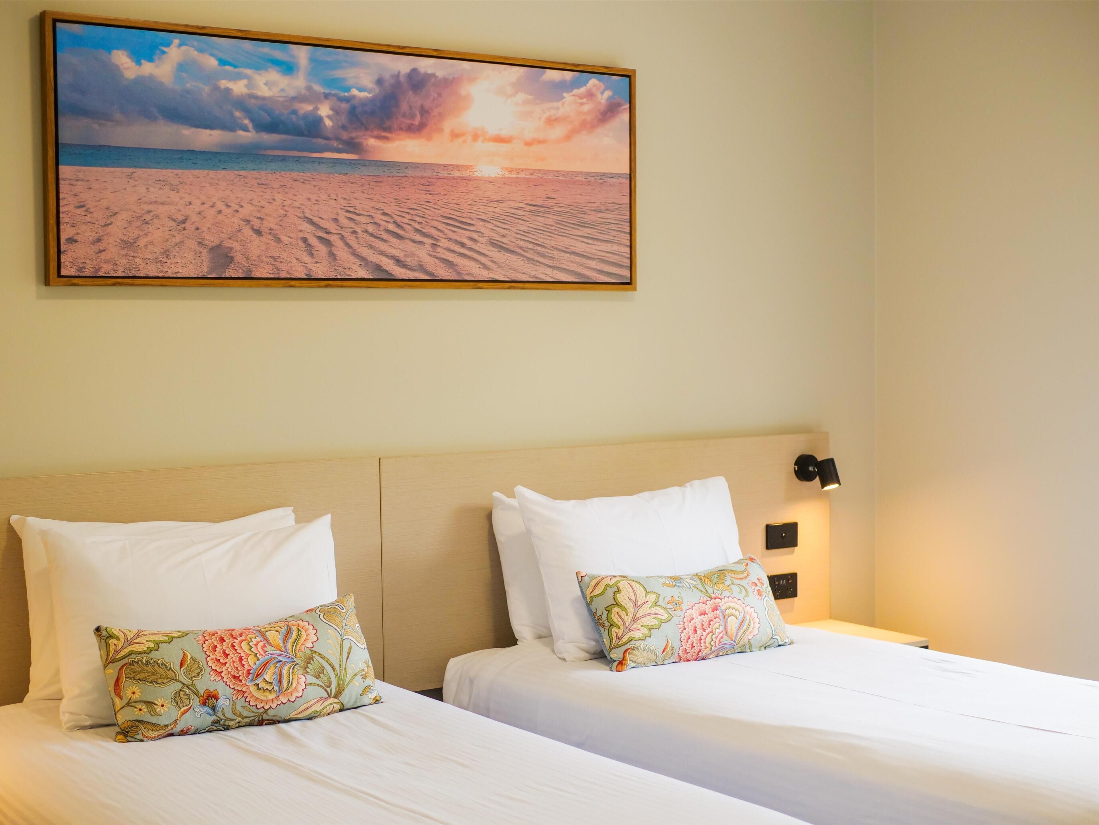 A hotel room with two beds, a large painting of a beach scene, and a night light on the wall.
