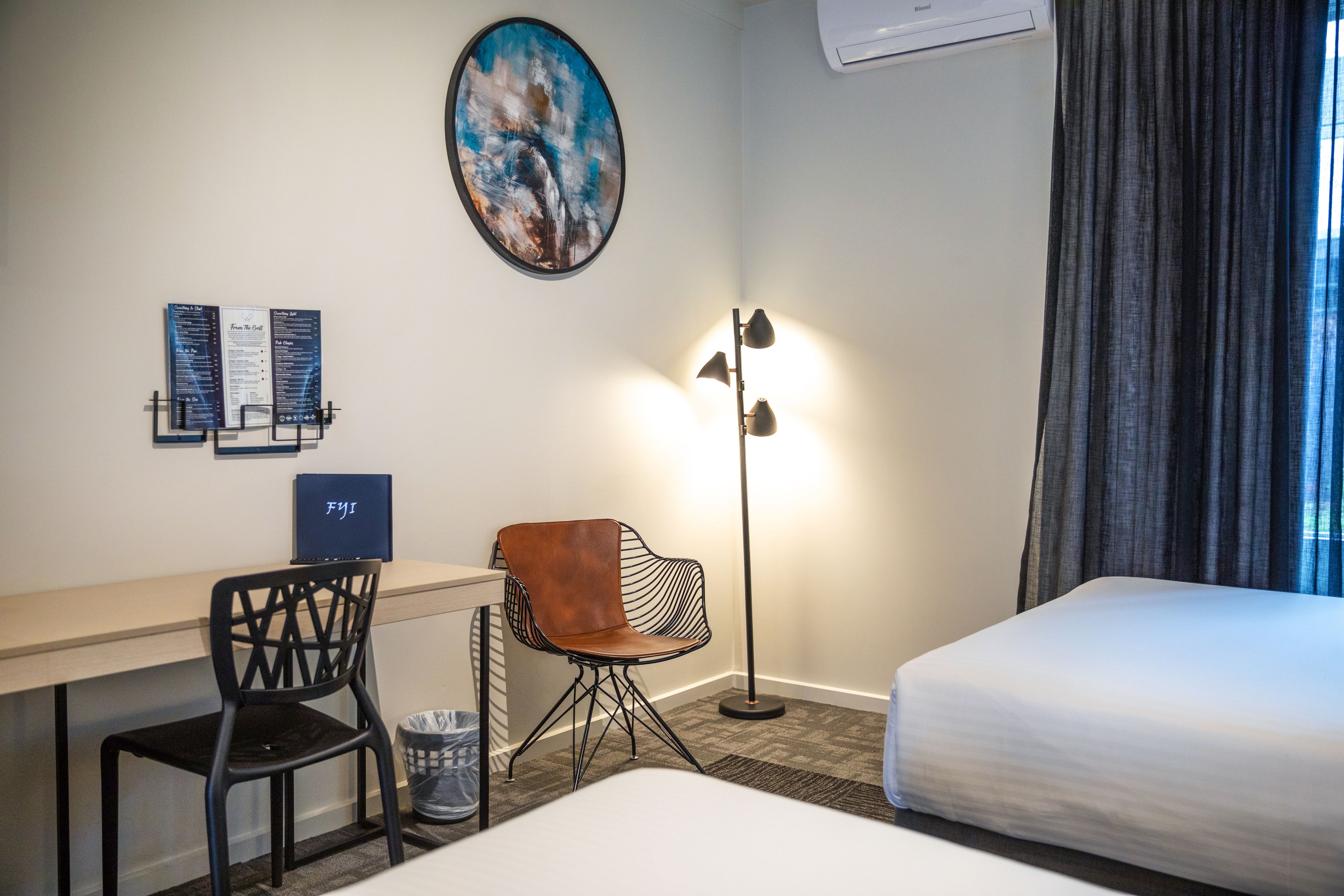 A hotel room with a desk, two chairs, a bed, a floor lamp, a painting, and a menu on the wall.