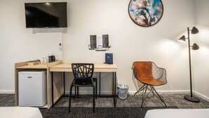 Et hotelværelse med en seng, et skrivebord med stol, et lille køleskab, et tv ophængt på væggen, et billede, en gulvlampe og en skraldespand.