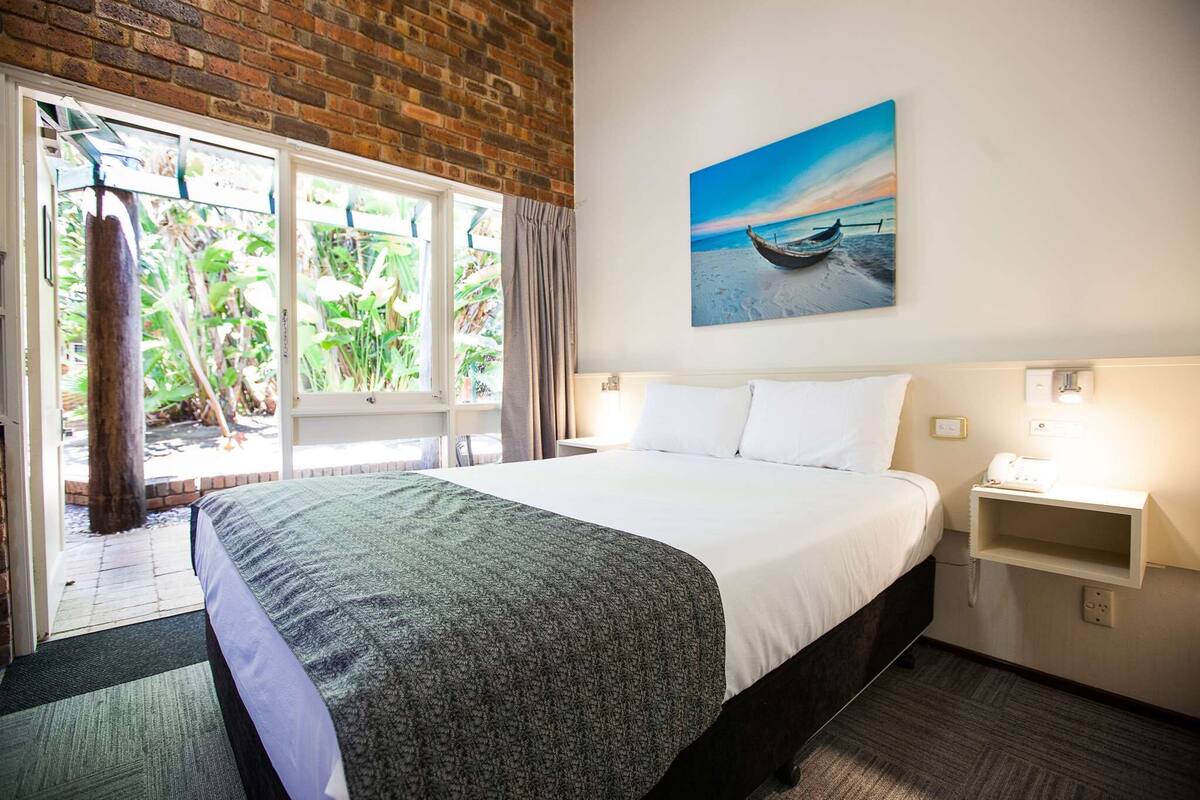 A hotel room with a large bed, a window with a view of greenery, a painting of a beach scene, and a bedside table with a phone.