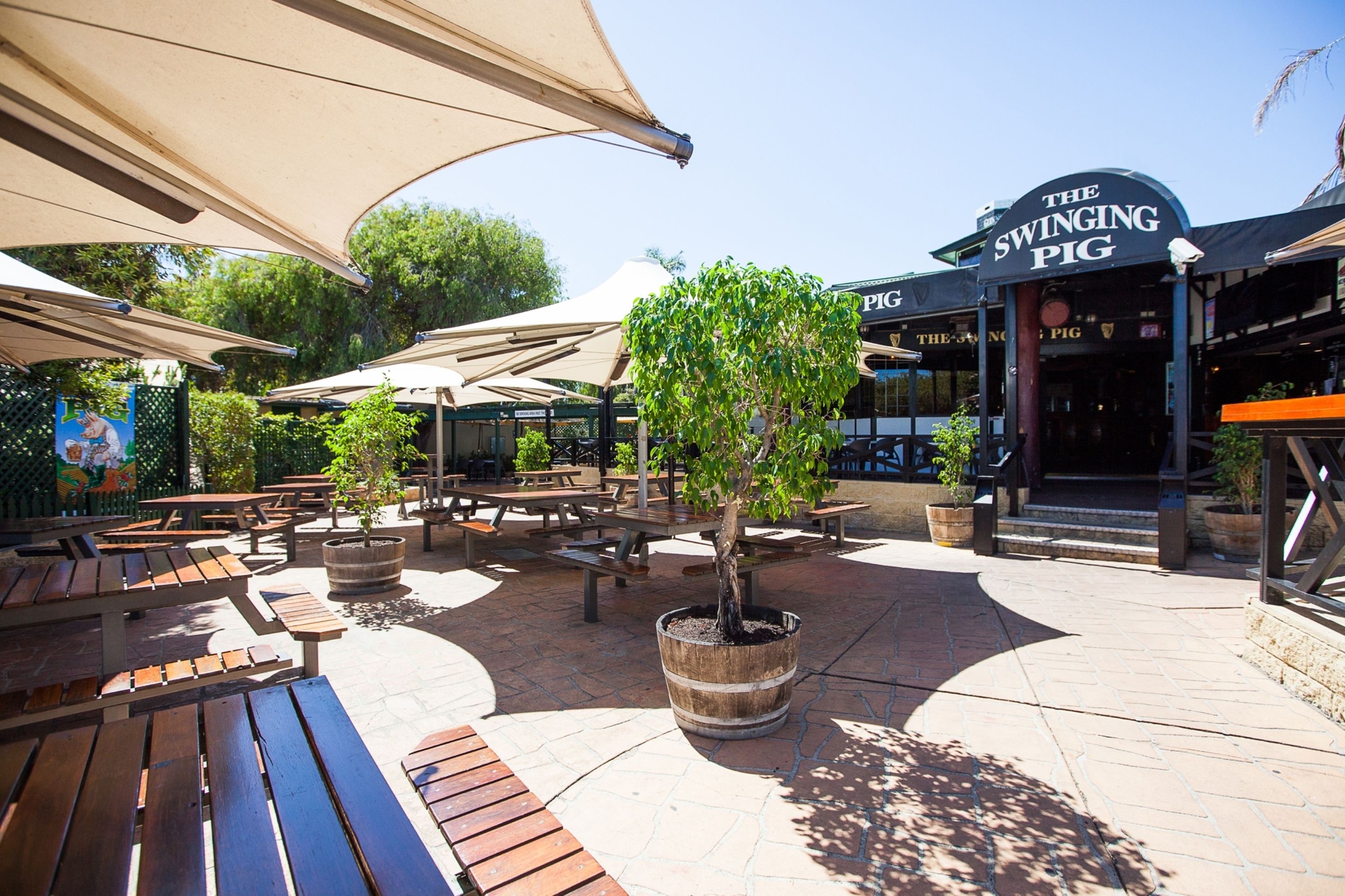 An outdoor patio area with wooden benches and potted trees, a sign for 'The Swinging Pig' restaurant, and a brick pathway.