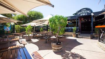 An outdoor patio area with wooden benches and potted trees, a sign for 'The Swinging Pig' restaurant, and a brick pathway.