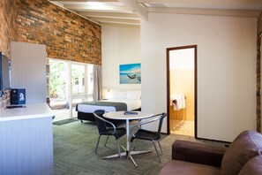 A hotel room with a brick wall, a bed, a desk with a computer, a round table with chairs, and a painting on the wall.