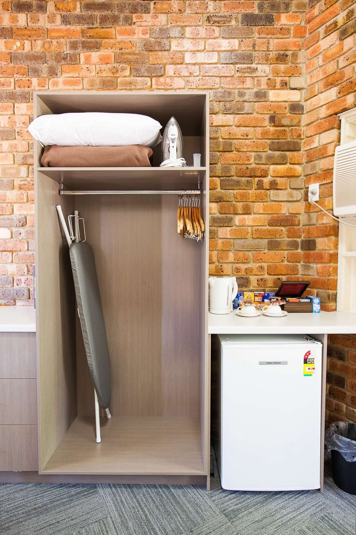 A compact room with a brick wall, a wardrobe with a hanging iron, a small refrigerator, and a shelf with kitchen items.