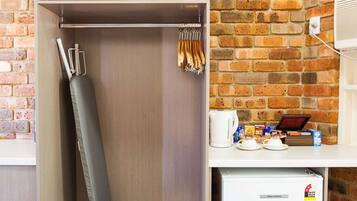 A compact room with a brick wall, a wardrobe with a hanging iron, a small refrigerator, and a shelf with kitchen items.