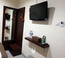 Habitación de hotel con un espejo grande, un televisor de pantalla plana montado en la pared, un estante pequeño con artículos de aseo y un cesto de basura.