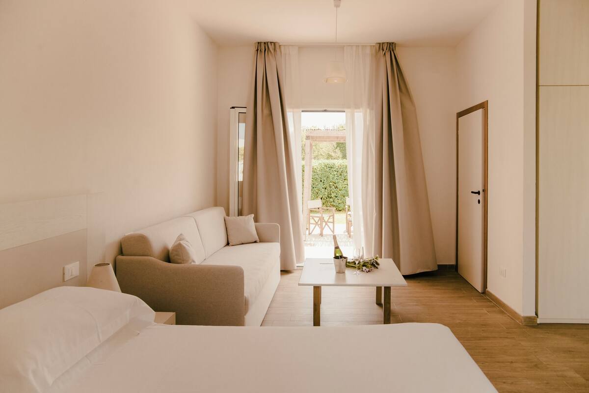 Una camera d'albergo con un letto, un divano, un tavolino con una bottiglia e fiori, e vista su un giardino attraverso una finestra.