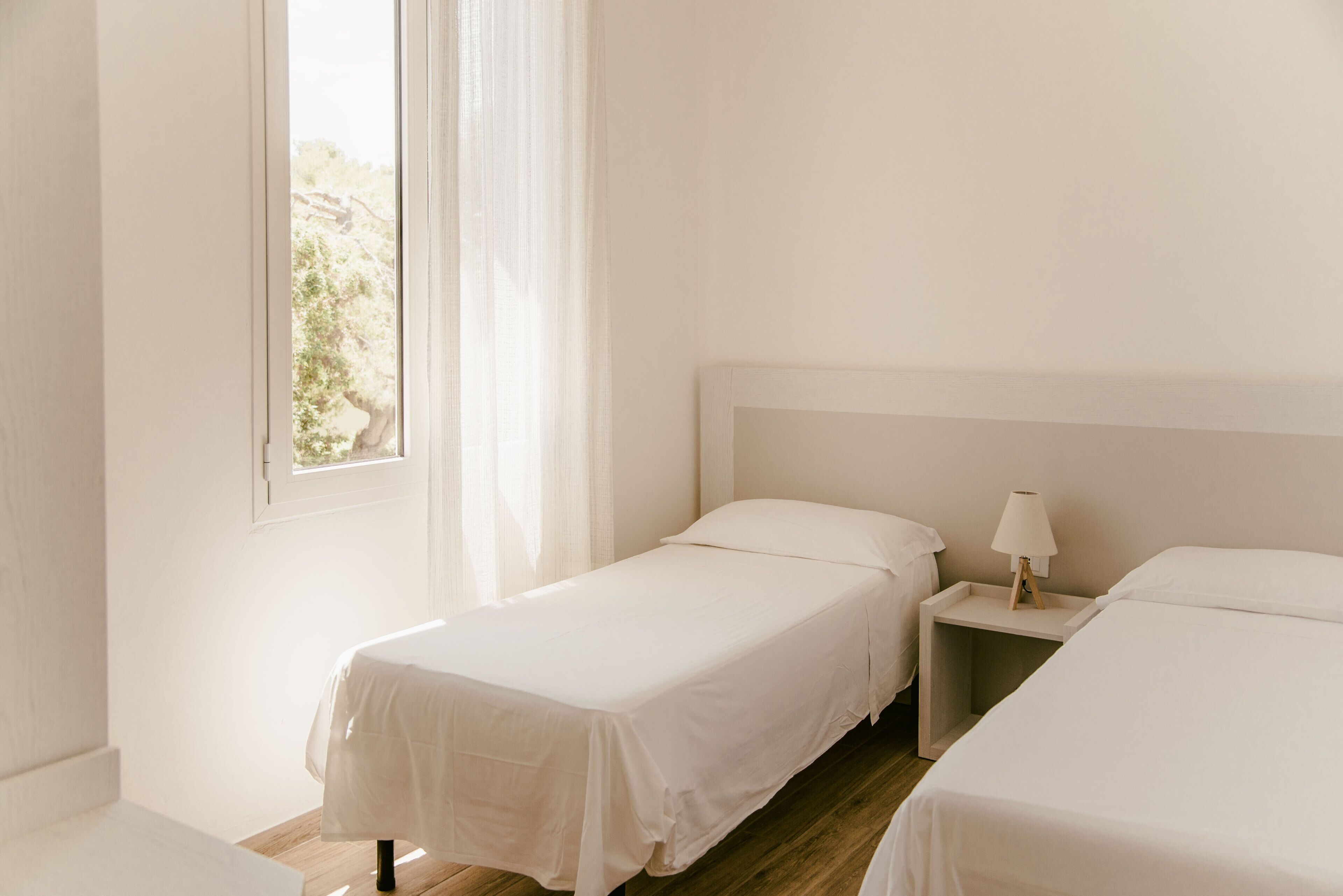 Ein helles, minimalistisches Schlafzimmer mit zwei Einzelbetten, einem Nachttisch mit Lampe und einem Fenster mit durchsichtigen Vorhängen.