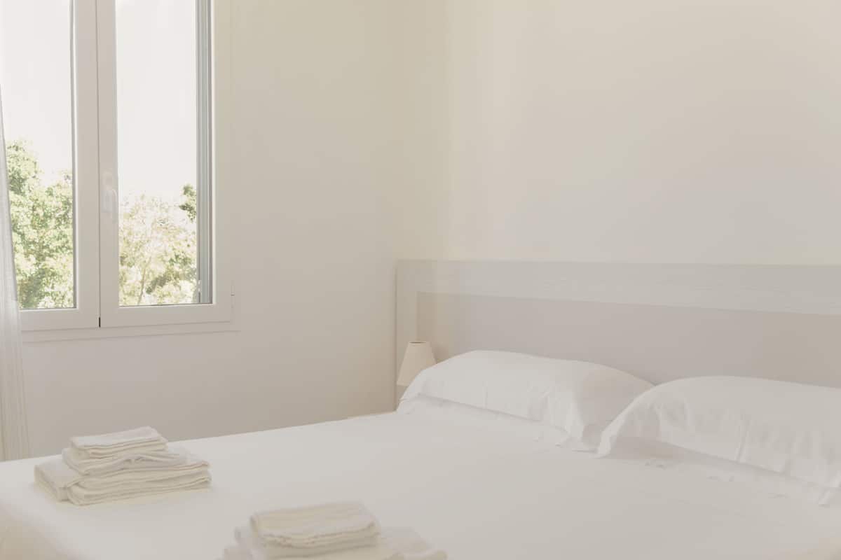 Camera da letto dal design minimalista con una grande finestra, un letto rifatto e un mucchio di asciugamani sul pavimento.
