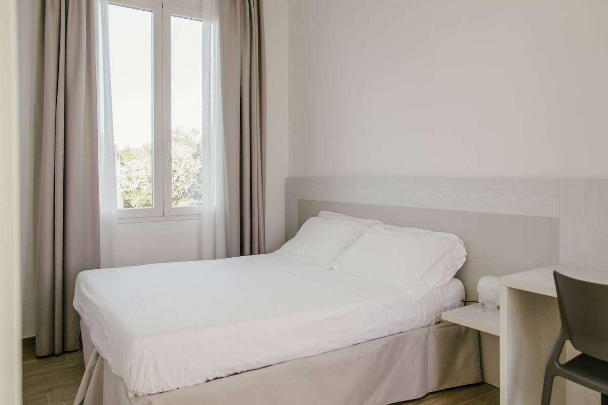 Un letto rifatto con lenzuola bianche, una piccola scrivania con una sedia e una finestra con tende.