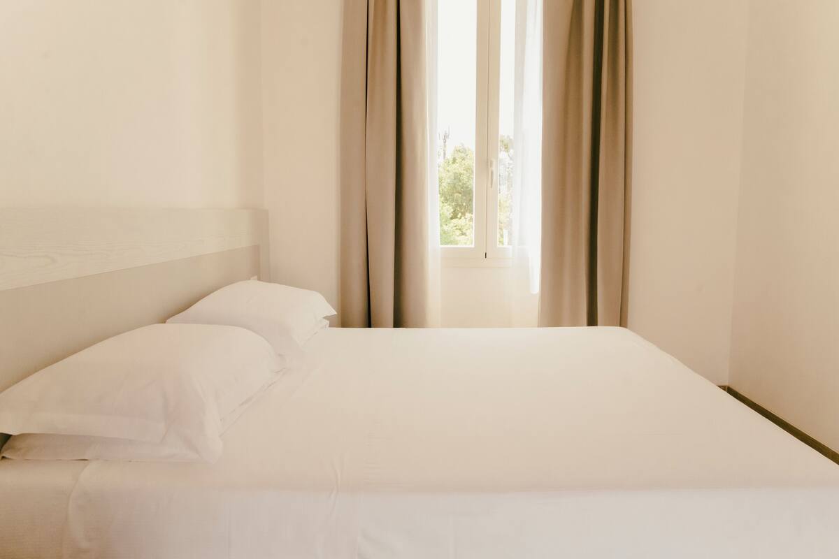 Un letto rifatto con lenzuola bianche in una stanza con un'ampia finestra e tende color beige.