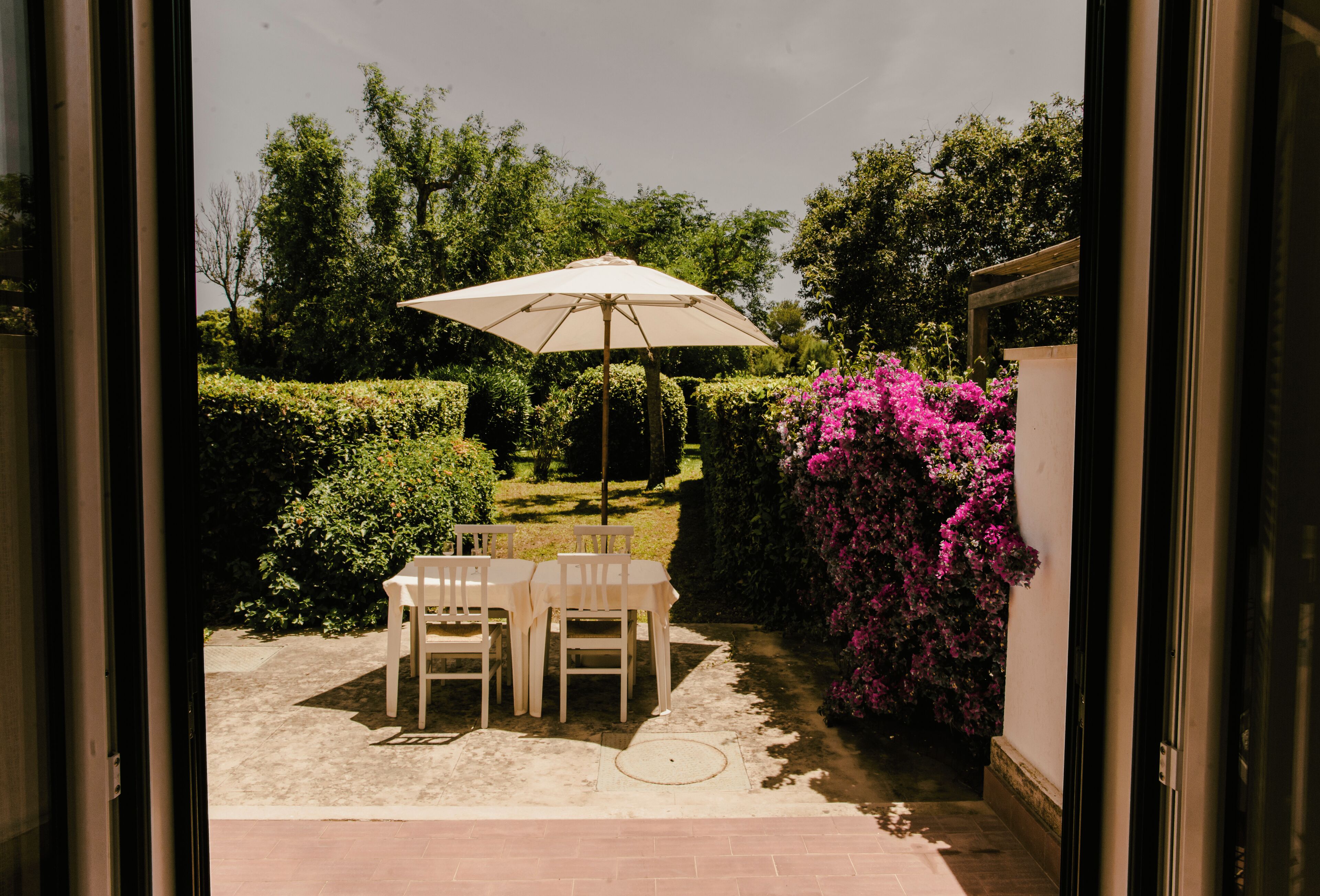 An outdoor patio with a table and chairs, a large umbrella, and a garden with bushes and flowers.