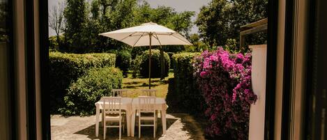 Un patio exterior con una mesa y sillas, un gran paraguas y un jardín con arbustos y flores.