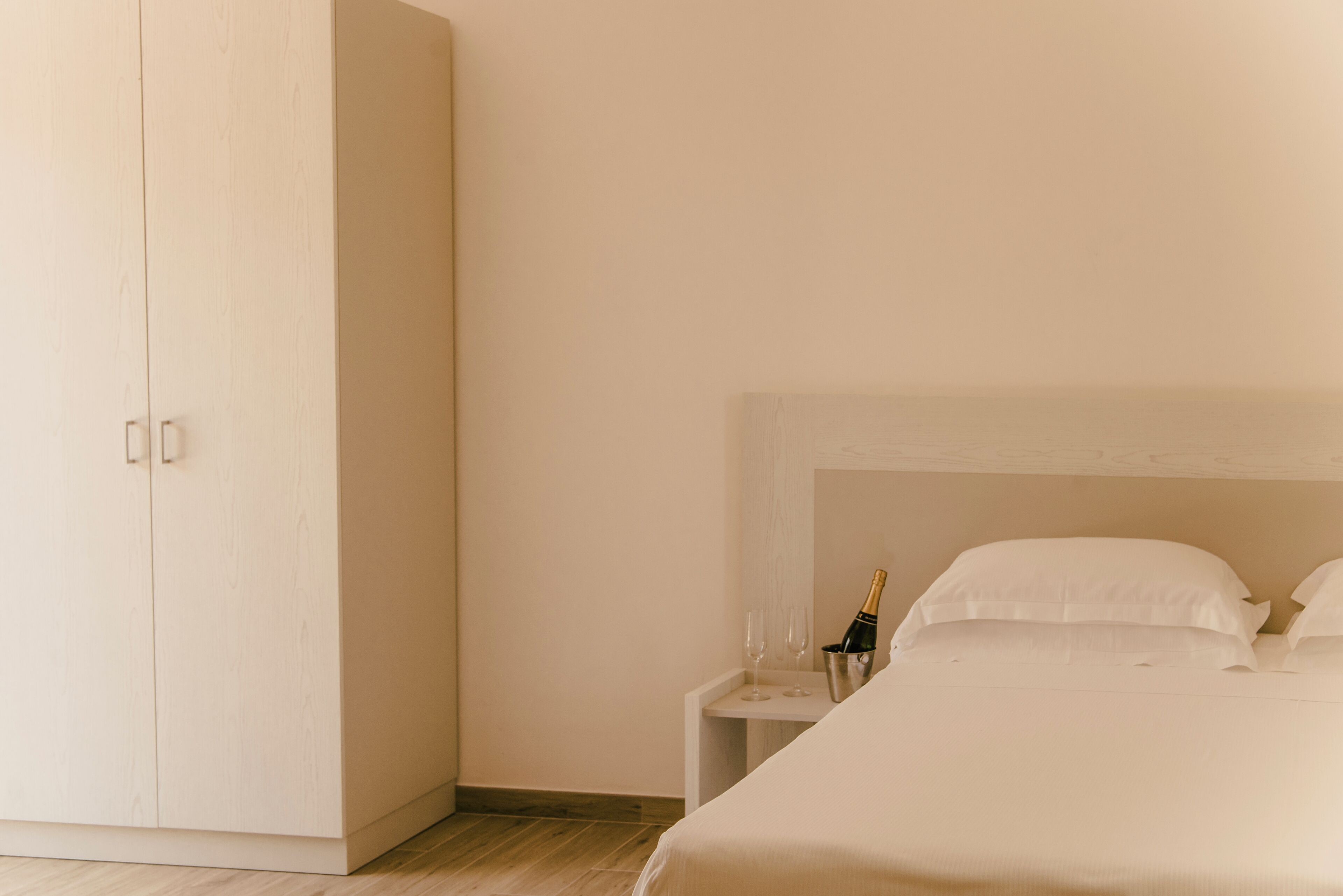 Habitación de hotel minimalista con un armario grande, una cama bien tendida y una mesita de noche pequeña con una botella de champán y copas.