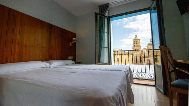 Habitación de hotel con una cama grande, cabecero de madera, balcón con vista a un edificio y una silla.