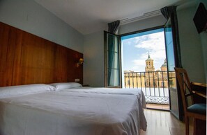 Habitación de hotel con una cama grande, cabecero de madera, balcón con vista a un edificio y una silla.