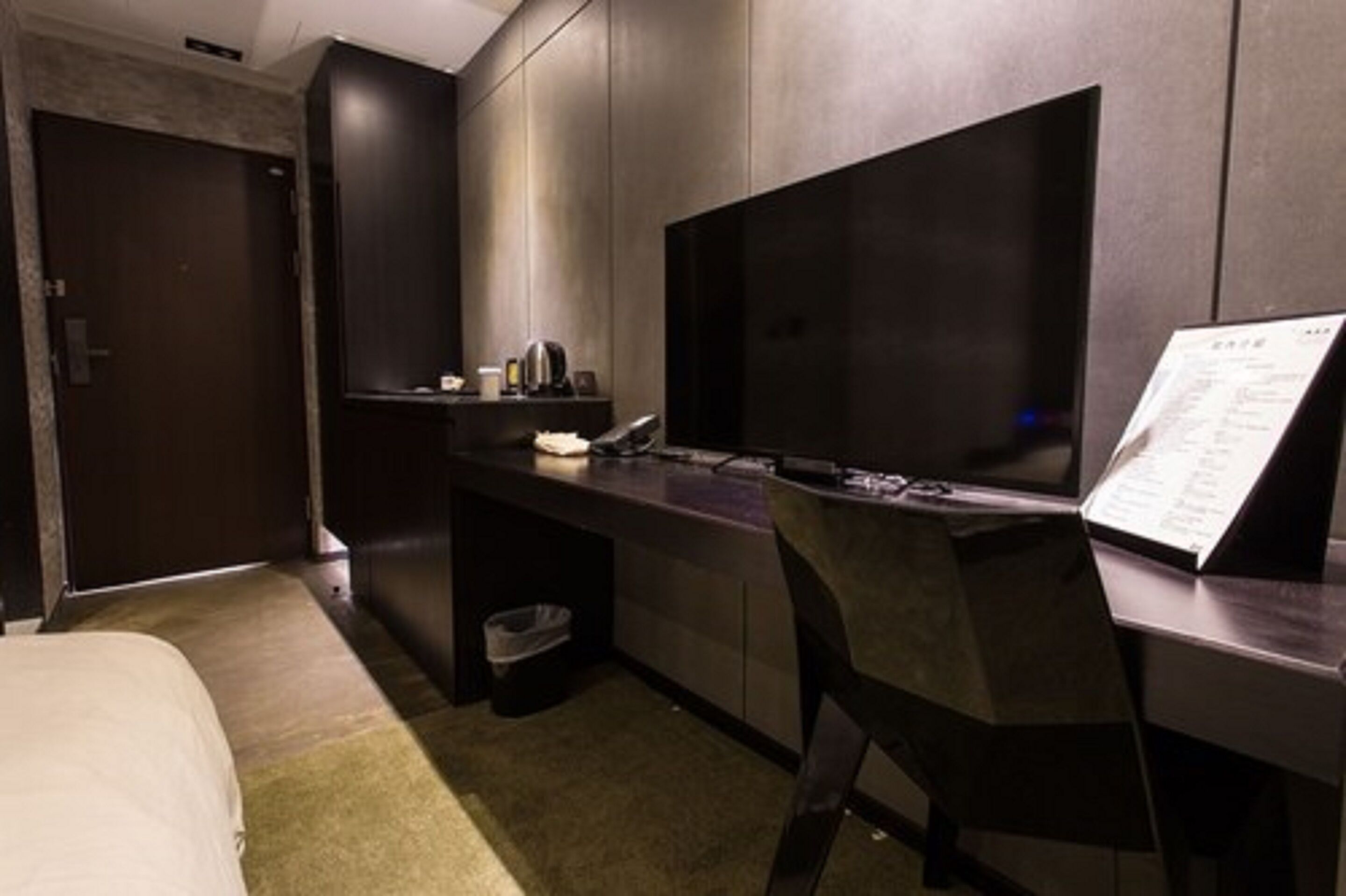 Een moderne hotelkamer met een groot bed, een bureau met een flatscreen televisie, een klein plankje met toiletartikelen en een prullenbak.