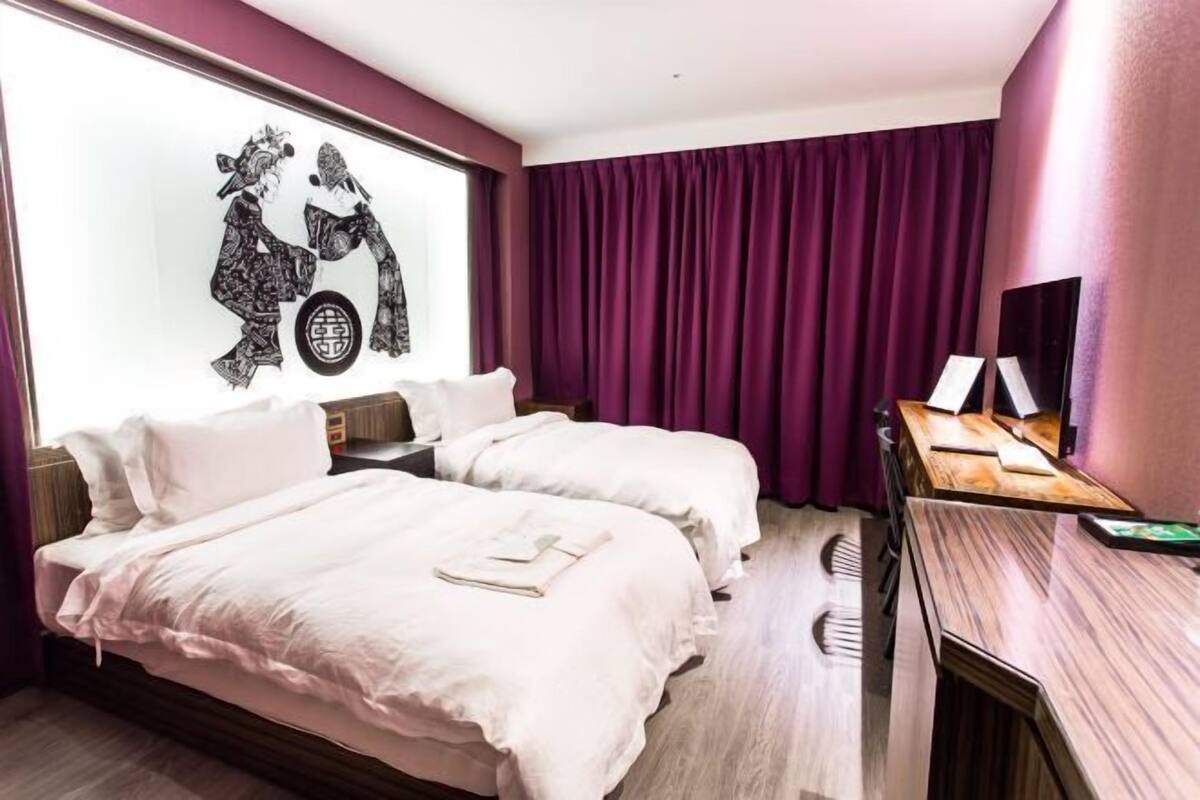 Una habitación de hotel con dos camas, un escritorio y un ventanal con cortinas moradas.