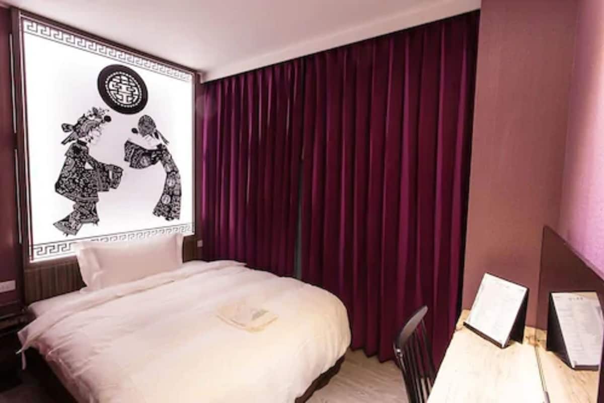 Una habitación de hotel con una cama, un escritorio con silla y un ventanal con cortinas.