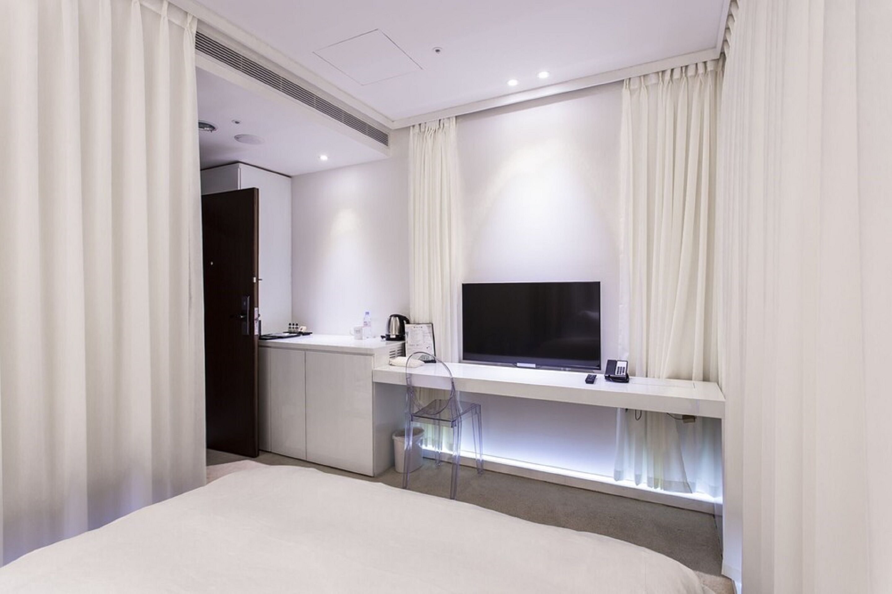 A modern hotel room with a bed, a desk with a TV, and a kitchenette.