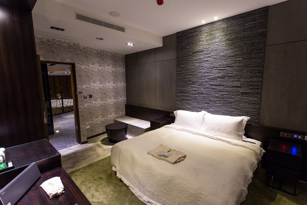 Quarto de hotel moderno com uma cama grande, uma pequena mesa de cabeceira, uma escrivaninha e um espaço de estar com uma cadeira.