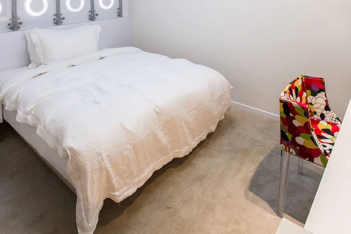 Un dormitorio con cama, una silla colorida, un aplique de techo y una pared con luces circulares.