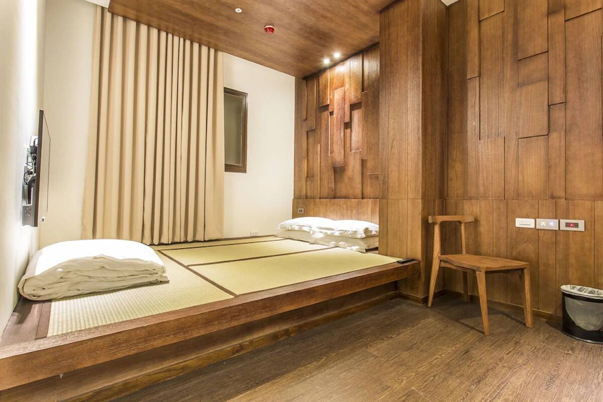 Una habitación tradicional japonesa con suelo de tatami, una cama baja de madera, una silla de madera y un pequeño cubo de basura.