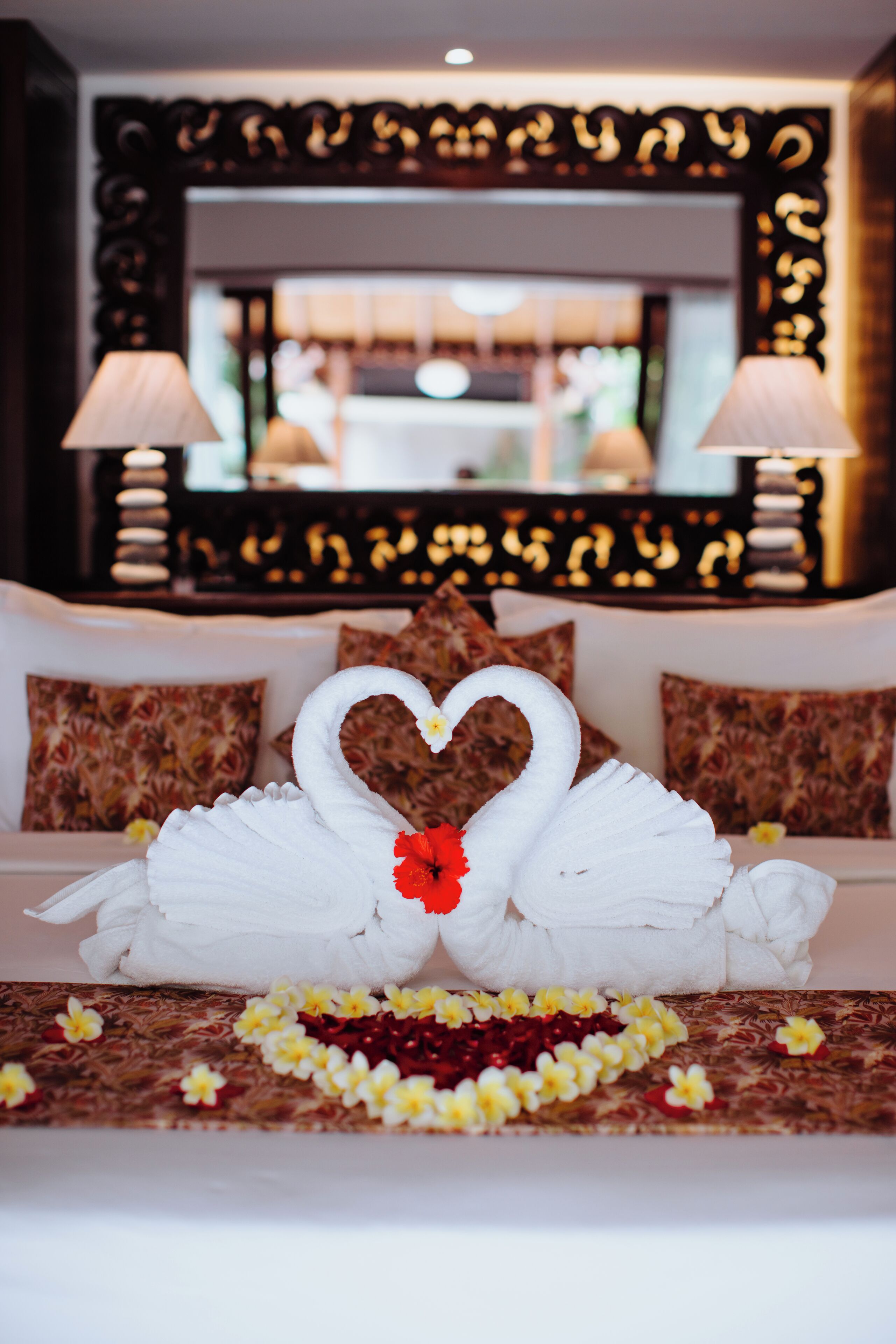 Una cama con una colcha con forma de cisne y un centro de mesa con forma de corazón, rodeada de arreglos florales.