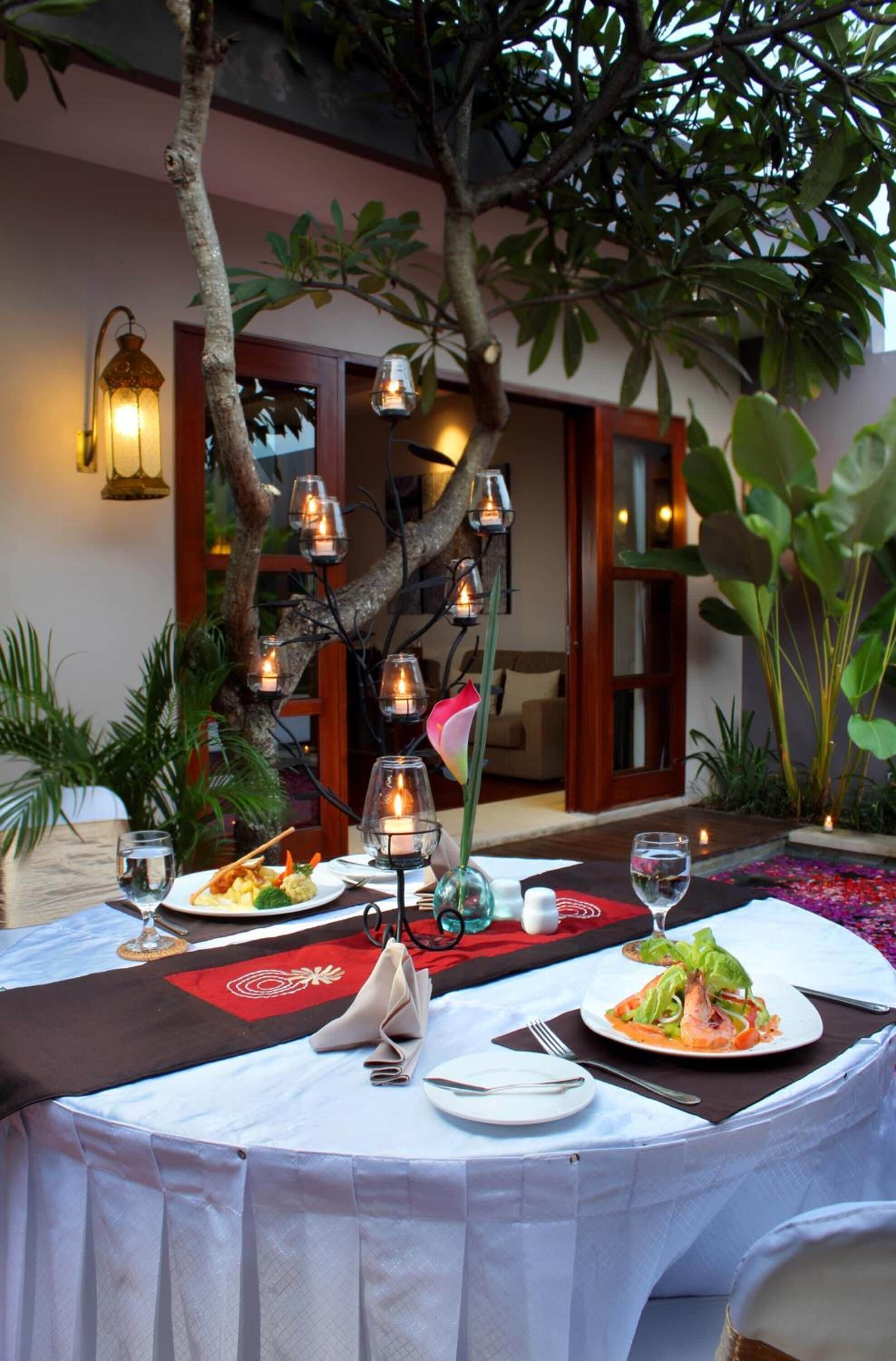 A dining table set outdoors with a tree in the center, surrounded by candles and a cozy atmosphere.