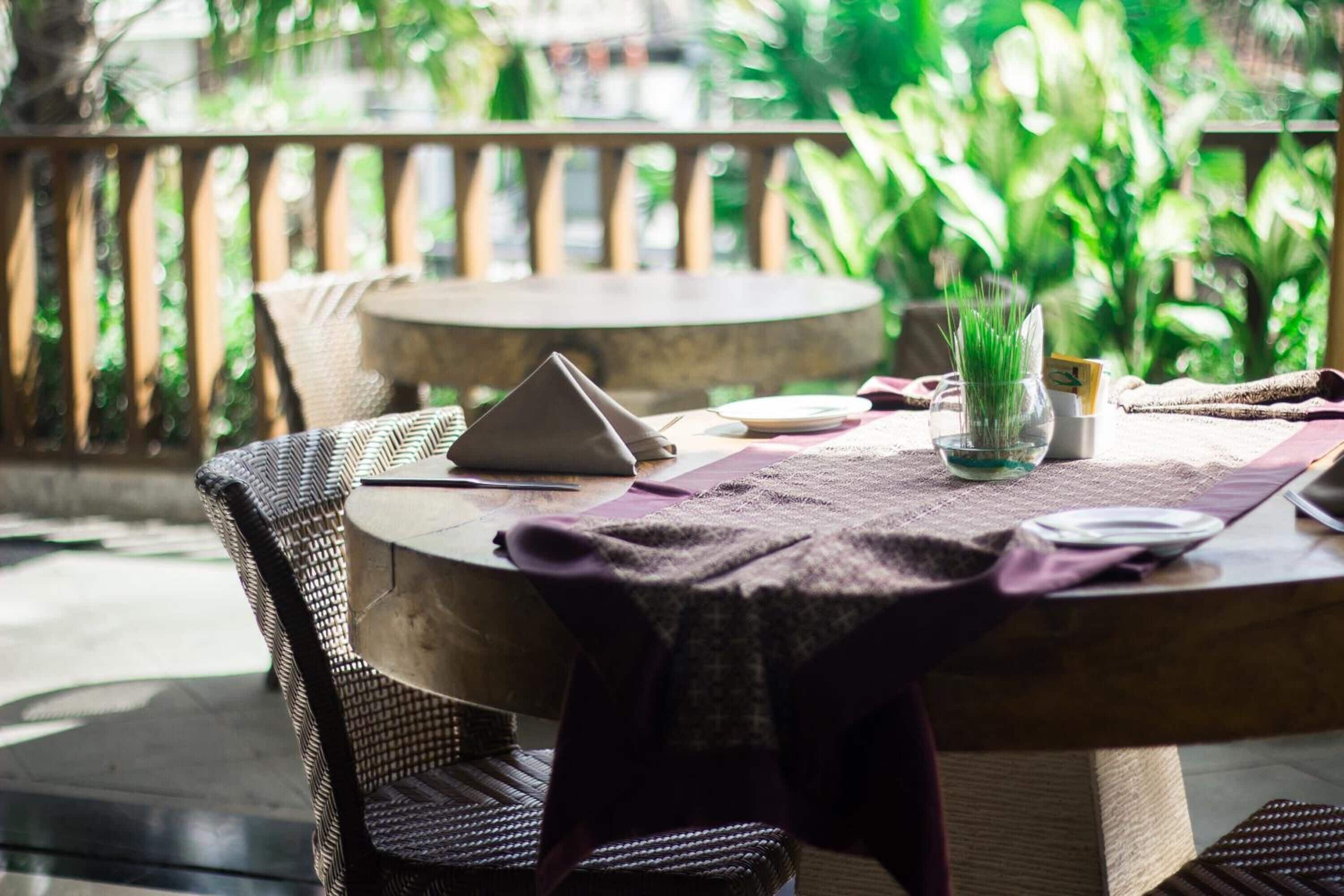 Una mesa redonda con una silla de mimbre, una computadora portátil, un florero de vidrio con plantas verdes y un mantel morado.