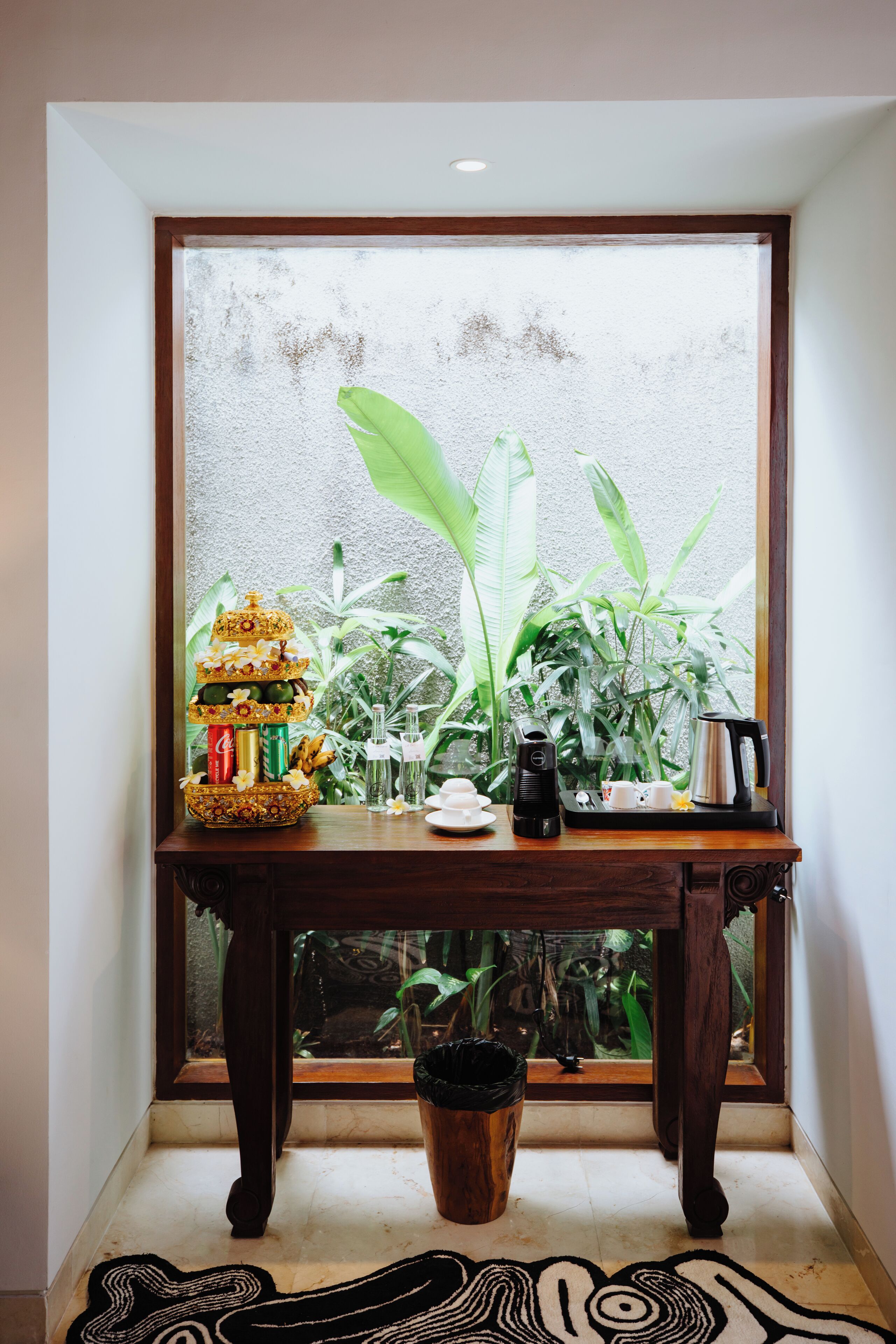 Une table en bois ornée d’un objet décoratif, avec une machine à café et une plante en pot, près d’une fenêtre donnant sur un paysage verdoyant.