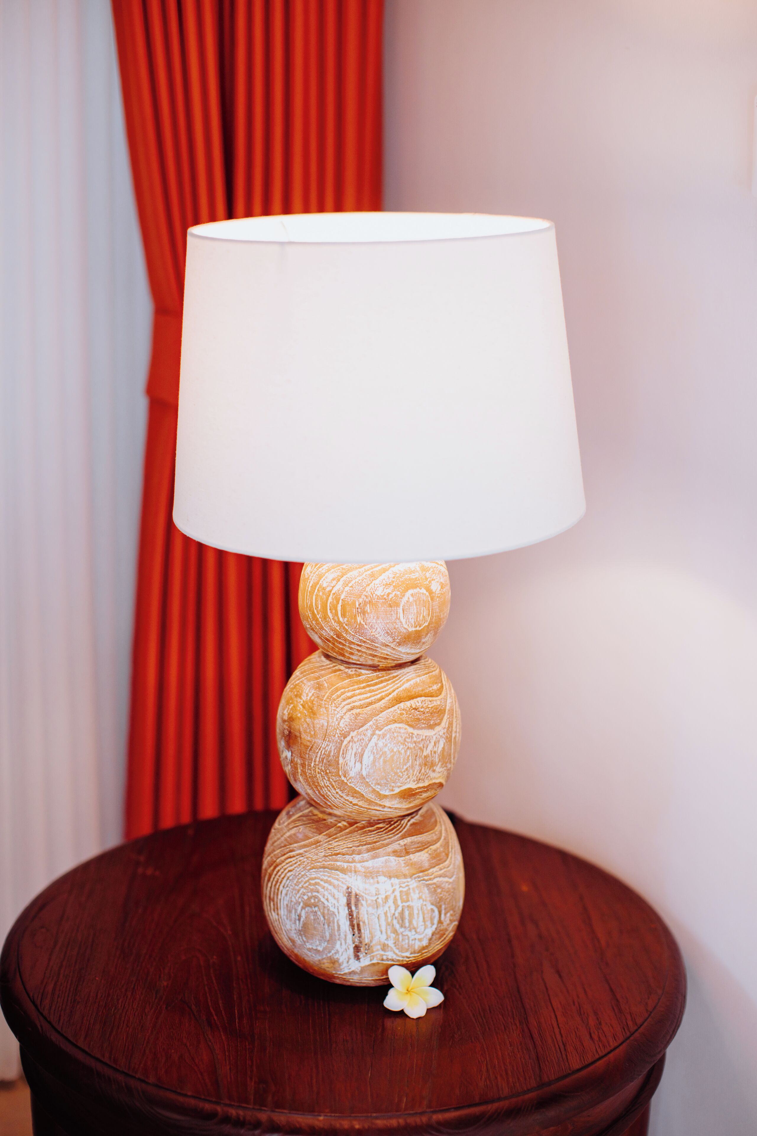 A wooden table lamp with a white shade on a round wooden table.