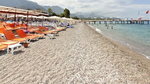 A pebble beach with sun loungers and umbrellas, a pier extending into the water, and mountains in the background. - Lims Bona Dea Beach Hotel – All Inclusive (Kemer)