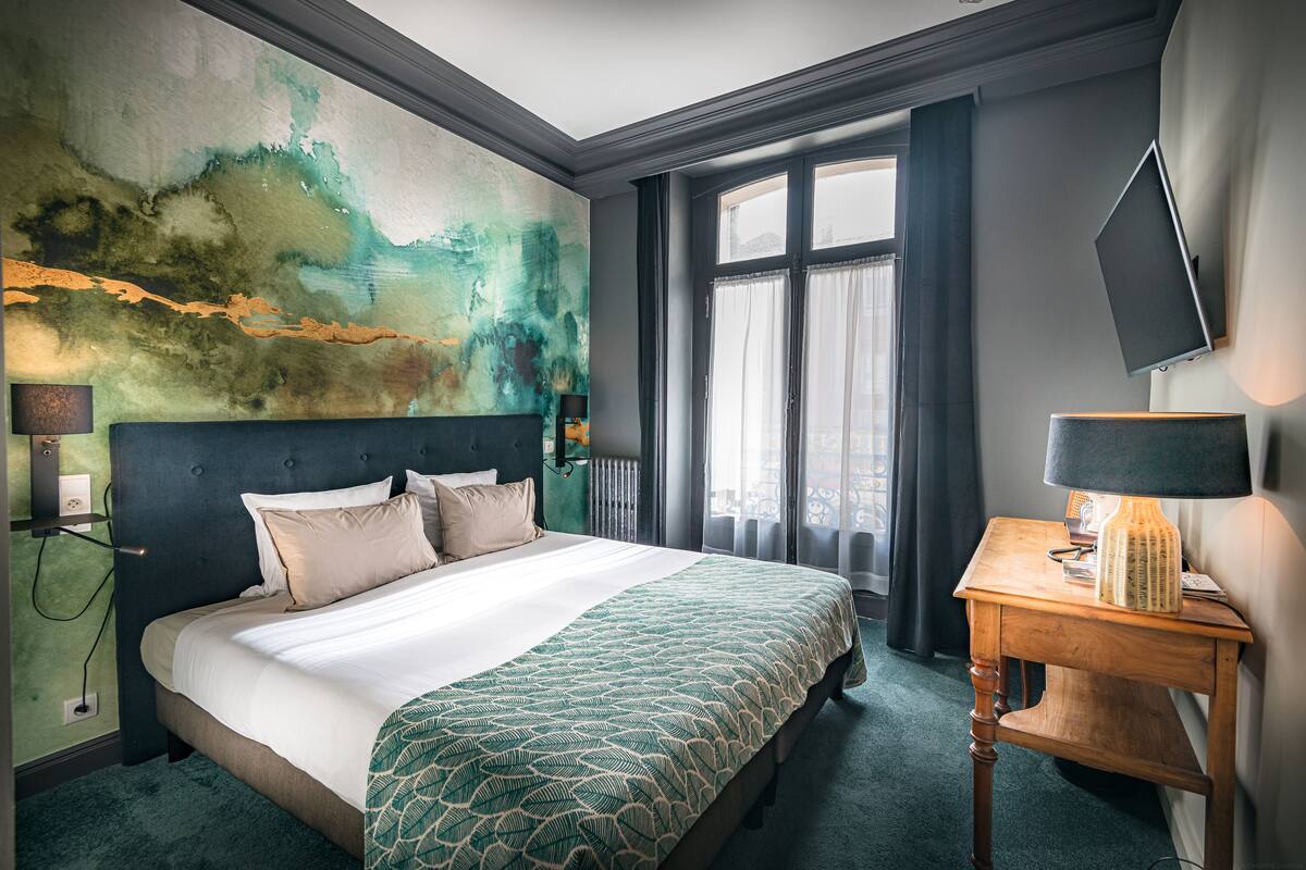Une chambre d’hôtel avec un grand lit, une table de chevet, un téléviseur fixé au mur et une fenêtre avec des rideaux.