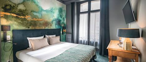 A hotel room with a large bed, a painting on the wall, a window with curtains, and a desk with a lamp.