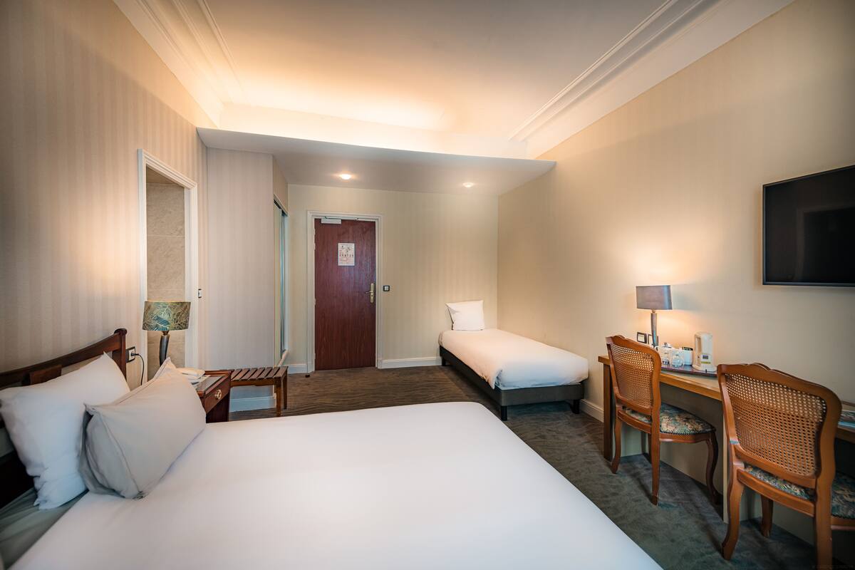 Une chambre d’hôtel avec un grand lit, un plus petit lit, un bureau avec des chaises, une télévision et une porte donnant sur une autre pièce.