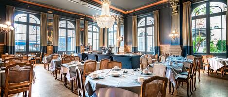 Un comedor amplio con múltiples mesas y sillas, ventanas grandes y candelabros ornamentados.