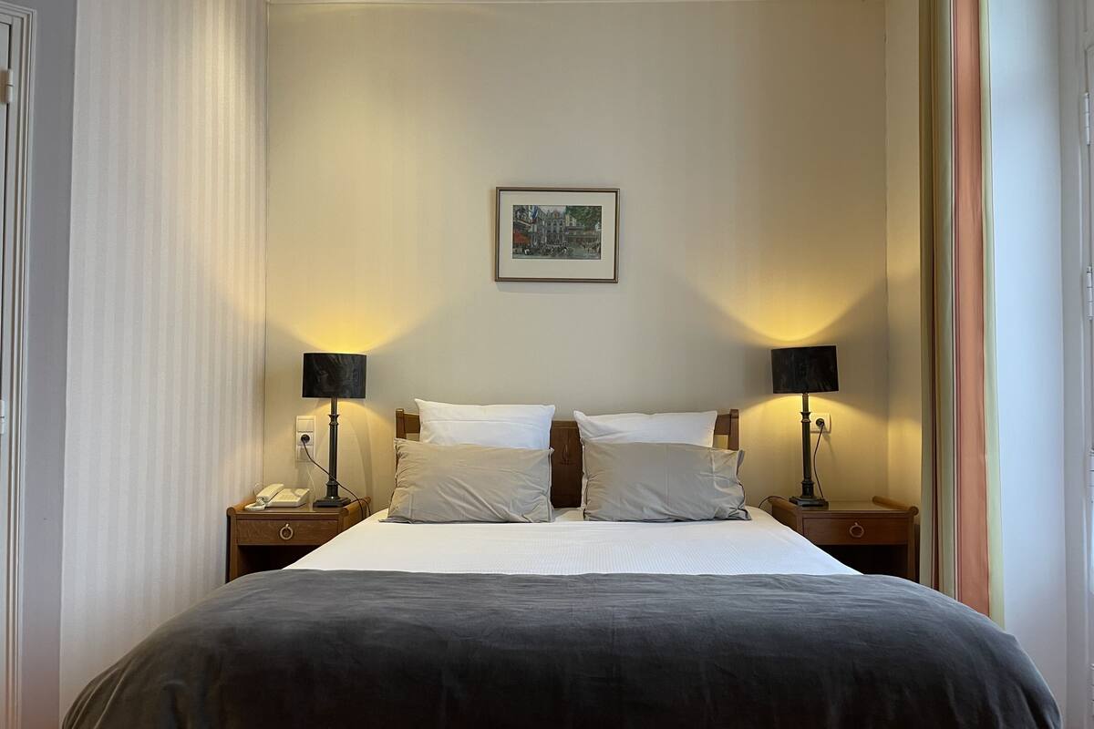 Une chambre d’hôtel avec un grand lit, deux tables de chevet, une petite table, une lampe, un tableau encadré au mur et une fenêtre avec des rideaux.