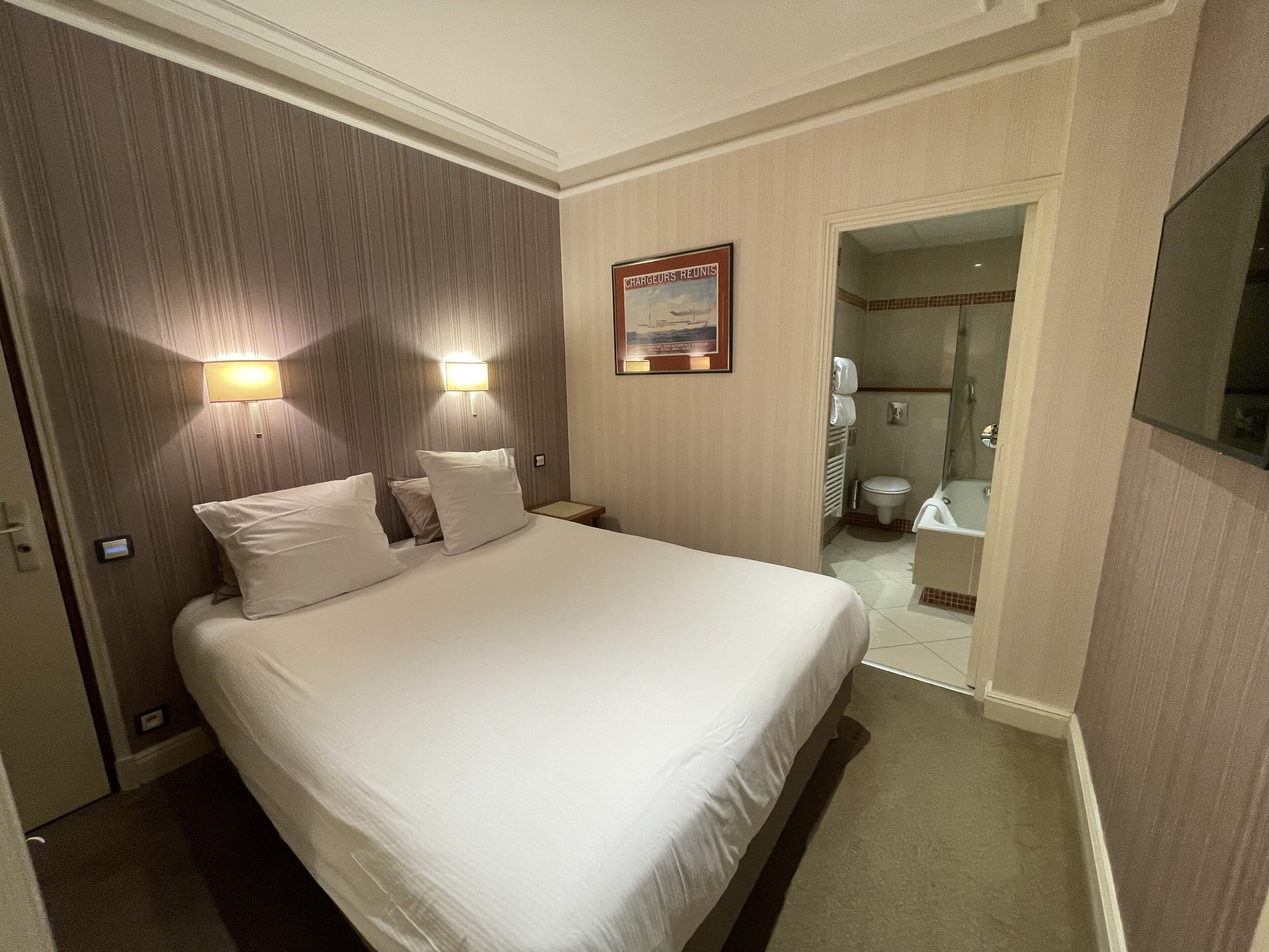 Una habitación de hotel con una cama grande, dos lámparas de noche, un cuadro en la pared y un baño visible a través de una puerta abierta.