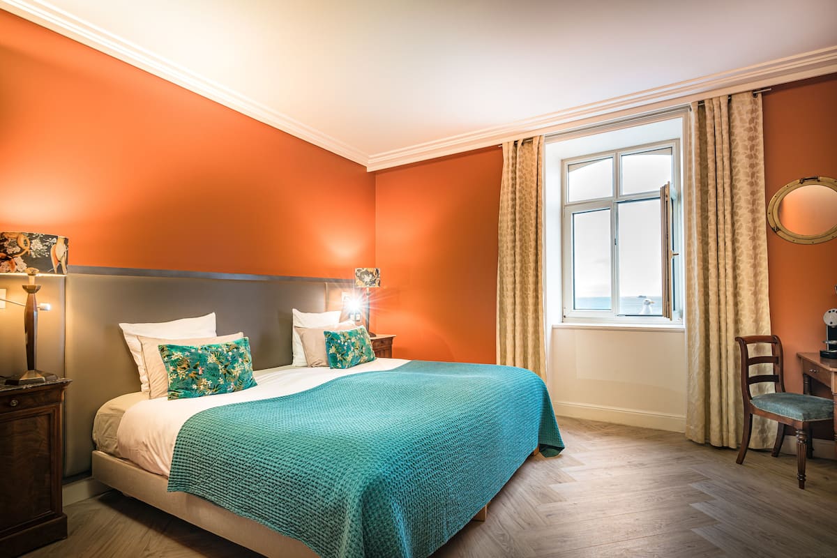 Une chambre avec un mur orange, un grand lit recouvert d’une couverture turquoise, deux oreillers, une table de chevet avec une lampe et une fenêtre avec des rideaux.