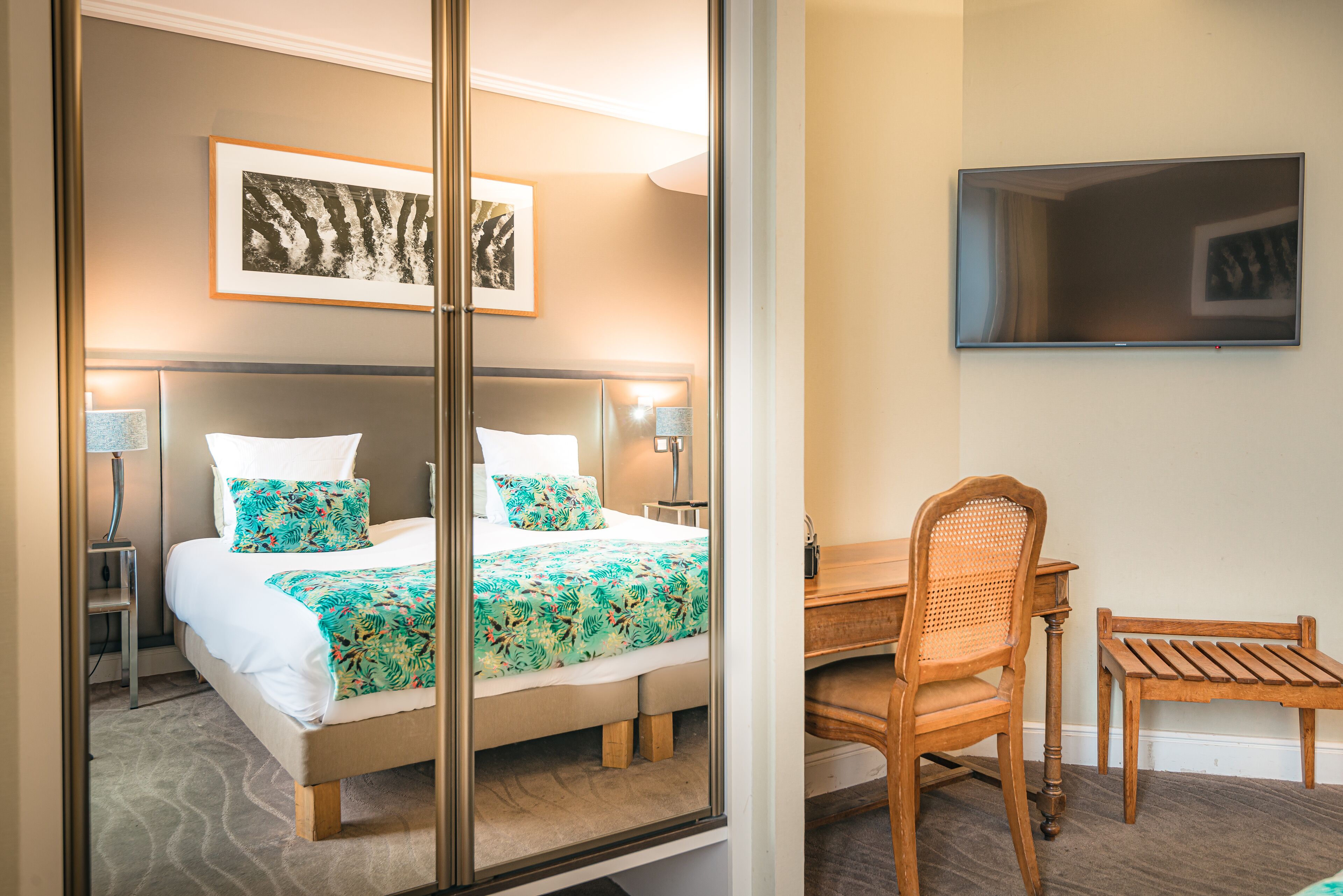 A hotel room with a bed, two bedside lamps, a desk with a chair, and a flat-screen TV on the wall.