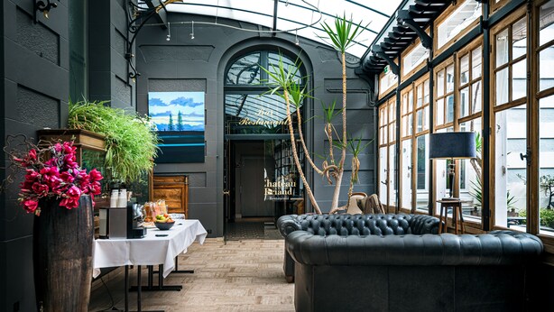 Interior de restaurante con piso de madera, un sofá de cuero negro, una mesa blanca con mantel blanco y un jarrón grande de flores rosas.