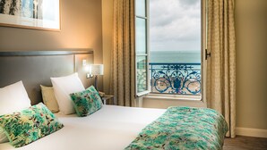 Habitación de hotel con cama, mesita de noche, lámpara, ventana con cortinas y vista al mar.