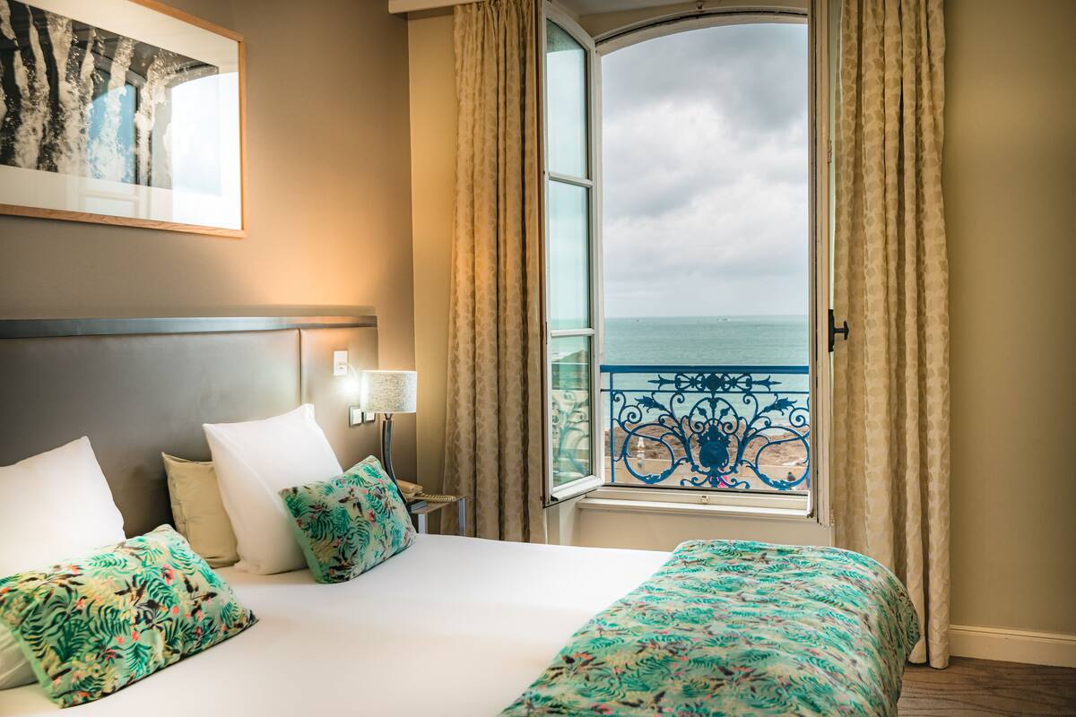 Une chambre d’hôtel avec un lit, une table de chevet, une lampe, une fenêtre avec des rideaux et une vue sur la mer.