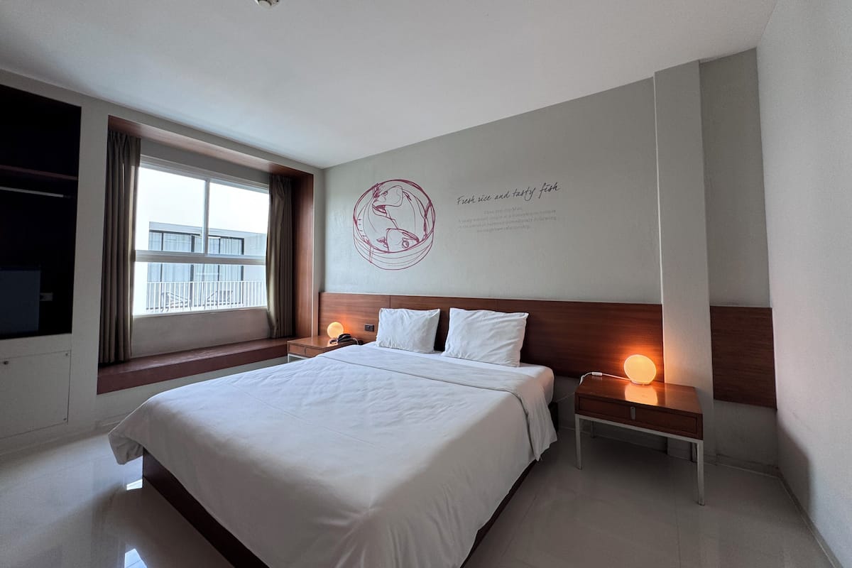 A hotel room with a large bed, a nightstand with a lamp, a window with curtains, and a wall with a circular design and text.