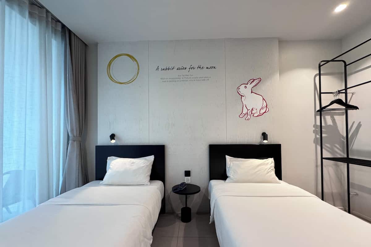 A hotel room with two beds, a wall-mounted artwork of a rabbit, and a small table with a lamp.