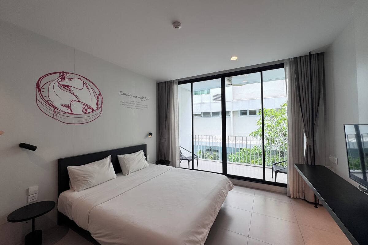 A modern hotel room with a large bed, a small bedside table, a flat-screen TV, and a balcony with a view of the city.