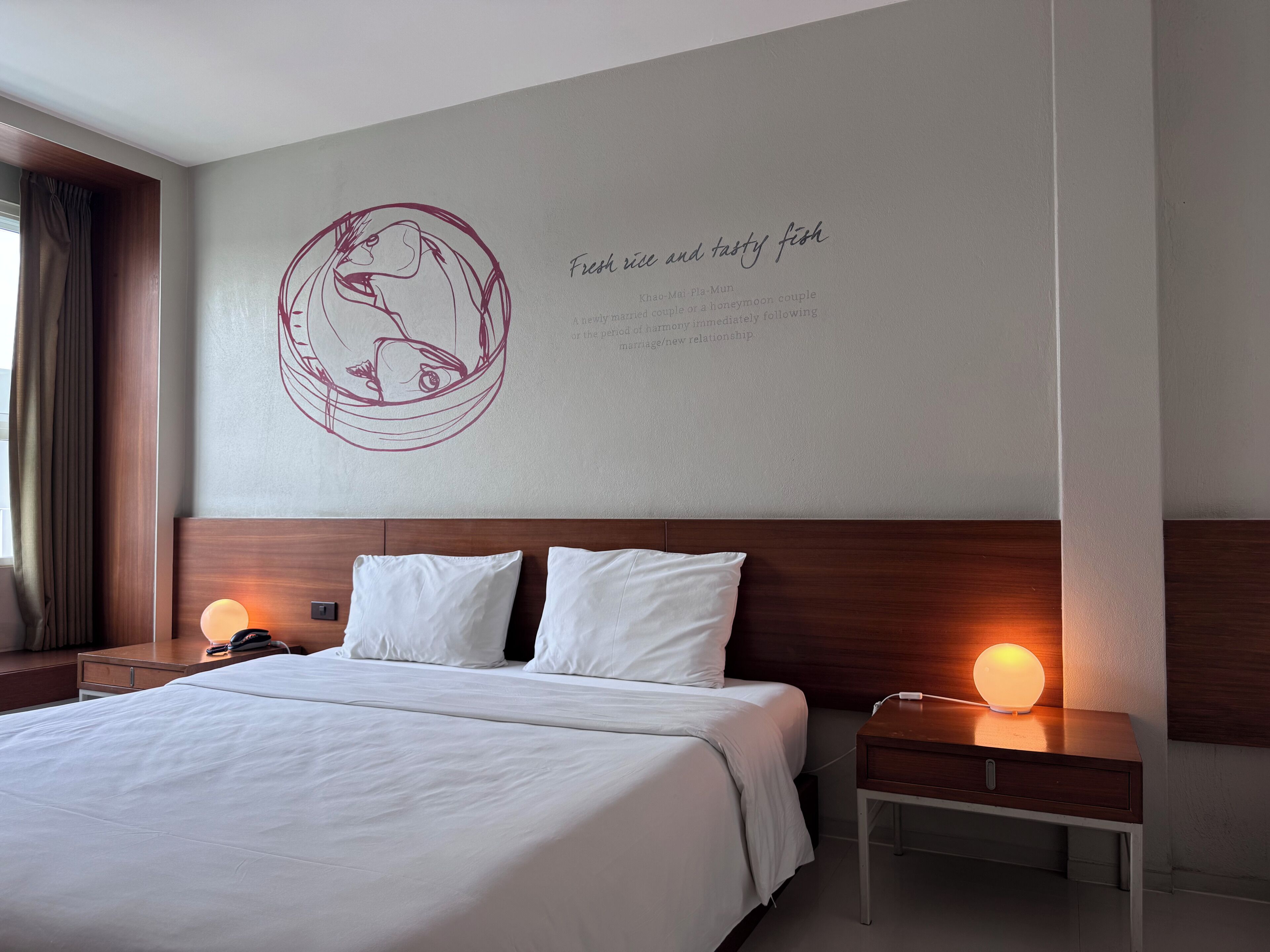 A hotel room with a large bed, two bedside tables with lamps, a wall with a circular design and text, and a window with curtains.